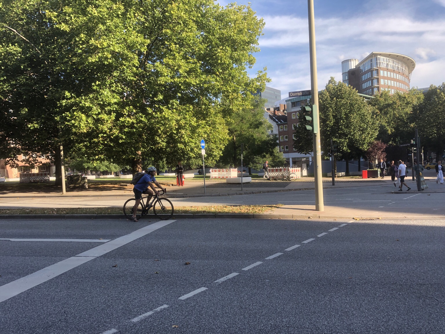 Ein banales Foto über eine Straße. Vorne eine Fahrradfahrer. Dahinter ein kleiner Park mit ein paar Baugatteren