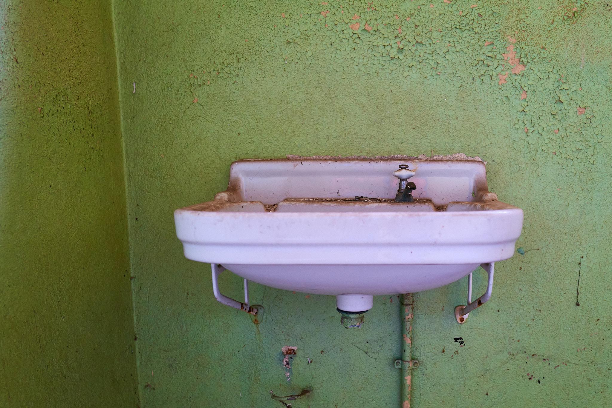 Ein altes, nicht mehr intaktes, Waschbecken an einer grünen Wand montiert. Die Farbe an den Wänden bröckelt stellenweise ab.