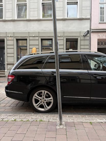 Ein Auto am Straßenrand, darüber ist im Hintergrund die Spitze eines Surfbretts und ein Ladenschild „Boardrider“ zu sehen. Die Schaufenster sind kaum sichtbar.