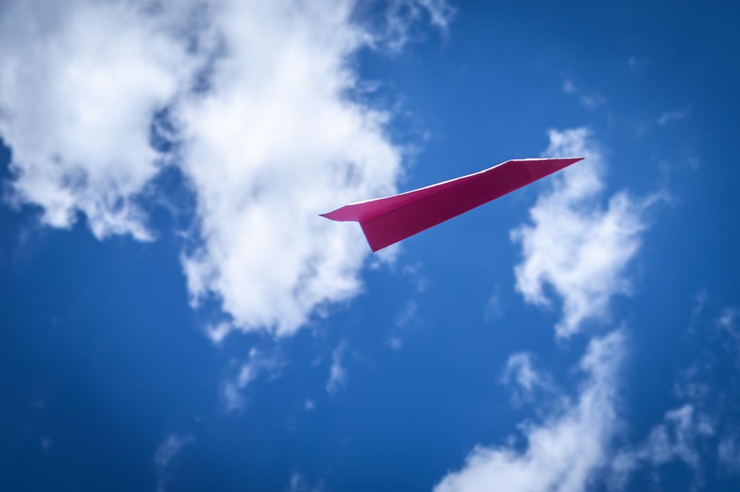 Rosaroter Papierflieger schwebt vor einem leuchtend blauen Himmel, umgeben von mehreren weissen Wolken.