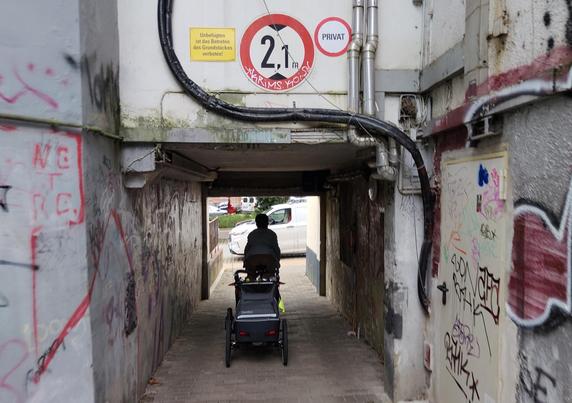 Jemand fährt mit Fahrrad und Anhänger aus einer Hinterhofeinfahrt auf die Straße. Auf der Auffahrt ist ein Auto geparkt.
