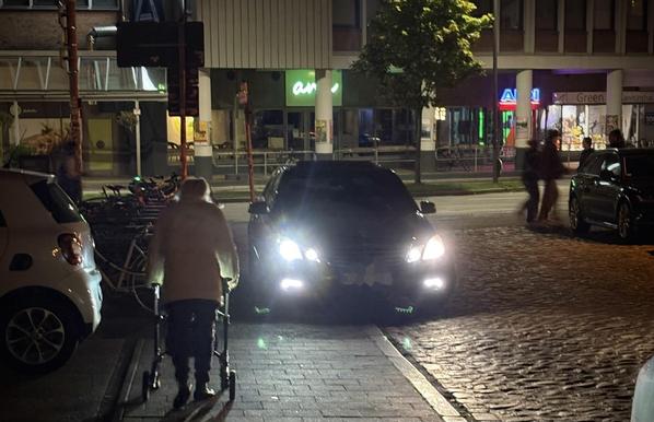 Eine Luxuslimousine steht auf einem Fußweg, zwischen Falschparker und Fahrradständern kommt kaum noch ein Mensch durch. Eine ältere Dame läuft mit Rollator auf die Engstelle zu.