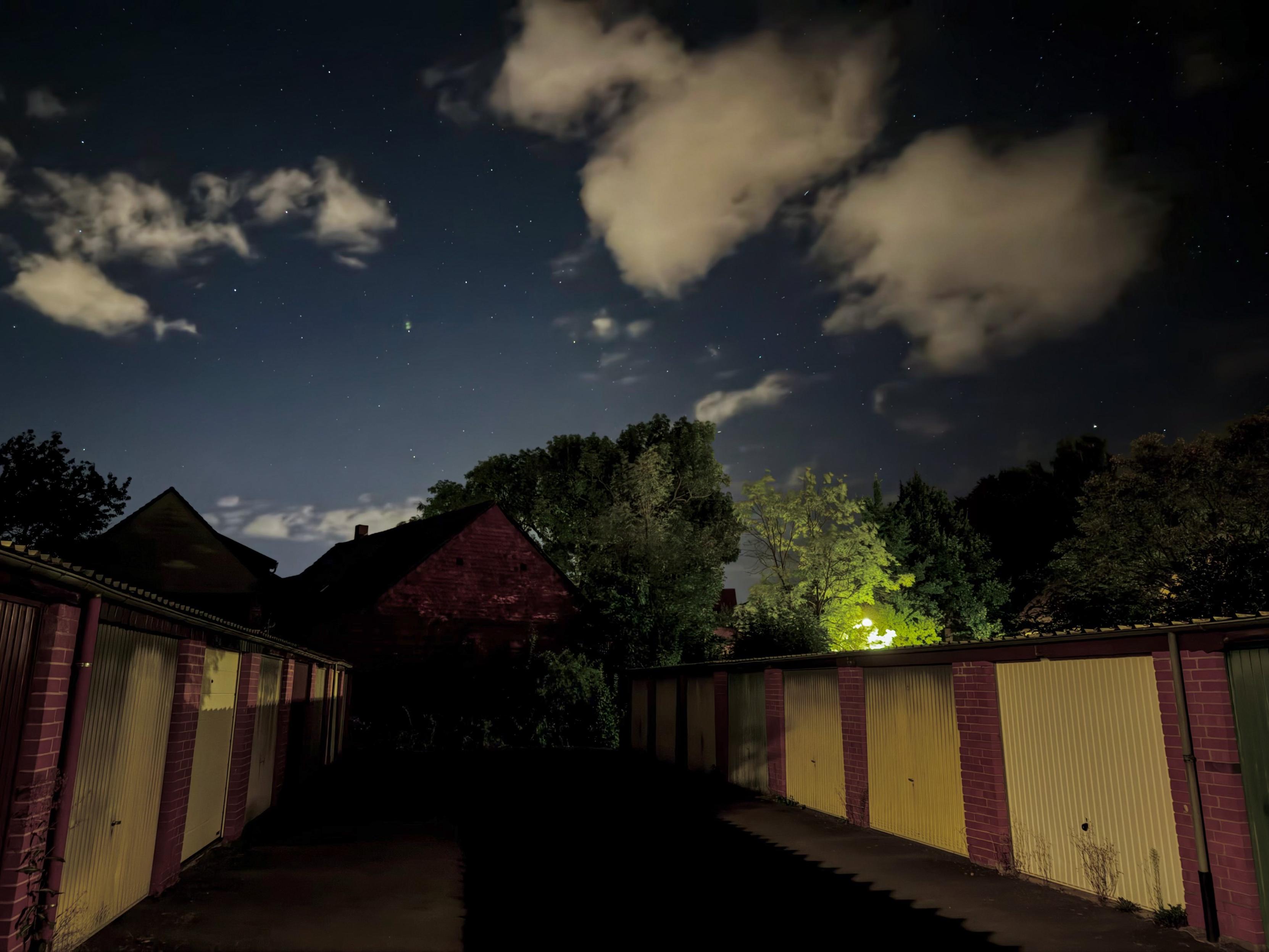 Eine Nachtszene mit einem dunklen Weg, der von Garagen gesäumt ist. Im Hintergrund sind unter einem Sternenhimmel mit vereinzelten Wolken die Silhouetten eines Hauses und von Bäumen zu sehen. Hinter den Bäumen strahlt ein warmes Licht, das der Szene einen zusätzlichen Kontrast verleiht.