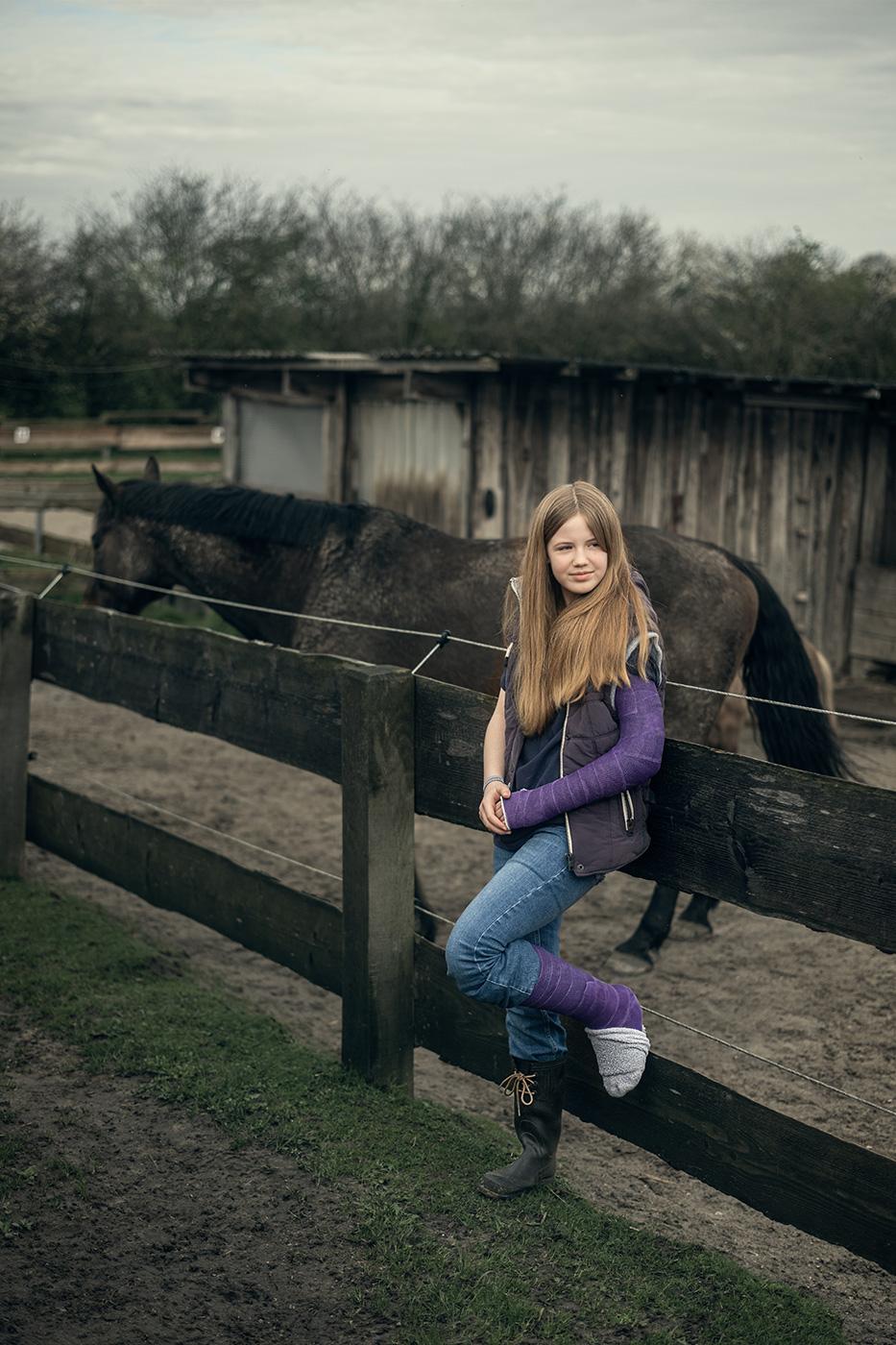 Ein 12-jähriges Mädchen mit langem, offenem Haar lehnt an einem Holzzaun. Ihr linkes Bein und ihr linker Arm sind in Gips, beide Gipse sind violett. Sie trägt eine blaue Jeans, eine dunkle Jacke mit lilafarbenen Ärmeln und einen schwarzen Gummistiefel. Die Zehen, die aus dem Gipsbein rausschauen würden, sind von einer grauen Socke verdeckt. Im Hintergrund steht ein dunkles Pferd vor einem hölzernen Stallgebäude. Die Szene spielt auf einem matschigen, ländlichen Gelände unter bewölktem Himmel.