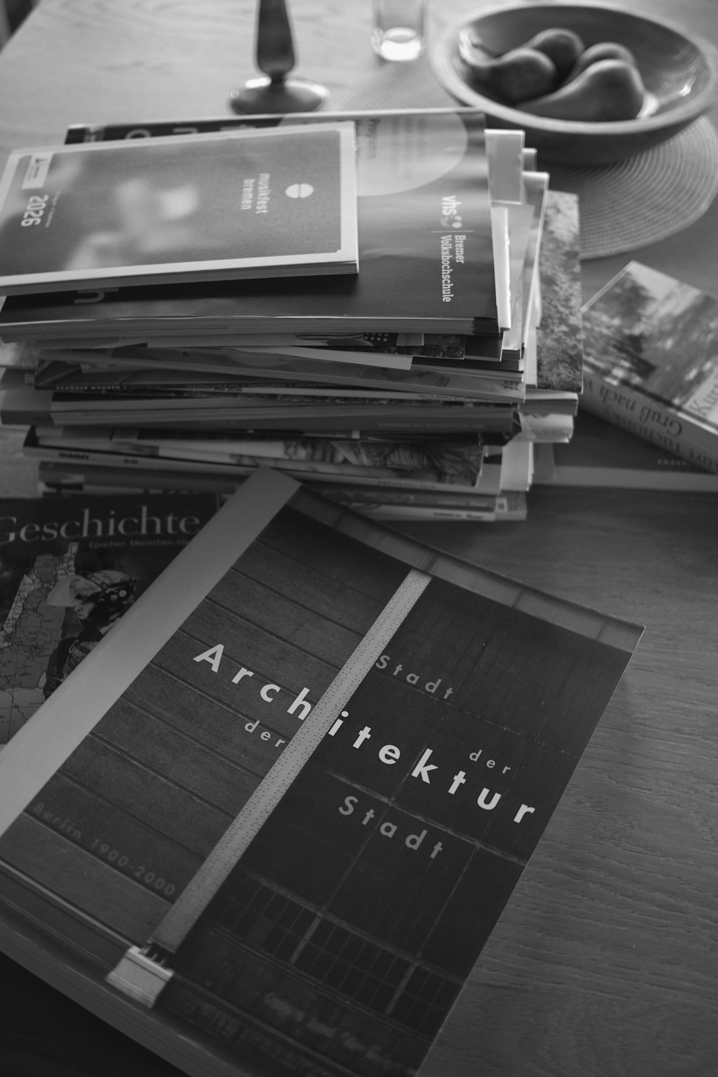 Schwarzweißaufnahme eines Stapels deutschsprachiger Architektur- und Kunstzeitschriften auf einem Holztisch. Im Vordergrund liegt das Magazin „Architektur der Stadt“ über Berliner Stadtarchitektur; darüber befindet sich unter anderem eine Ausgabe von „Bauwelt“ sowie weiteres Lesematerial. Im Hintergrund stehen ein Wasserglas, ein Löffel und eine Schale mit Obst.