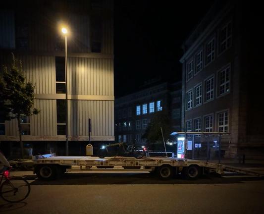 Ein großer Anhänger für den Transport von Baumaschinen steht auf einem Radweg, die Rampen zum Be- und Entladen sind angebracht. Auf dem Anhänger steht ein Lastenrad.