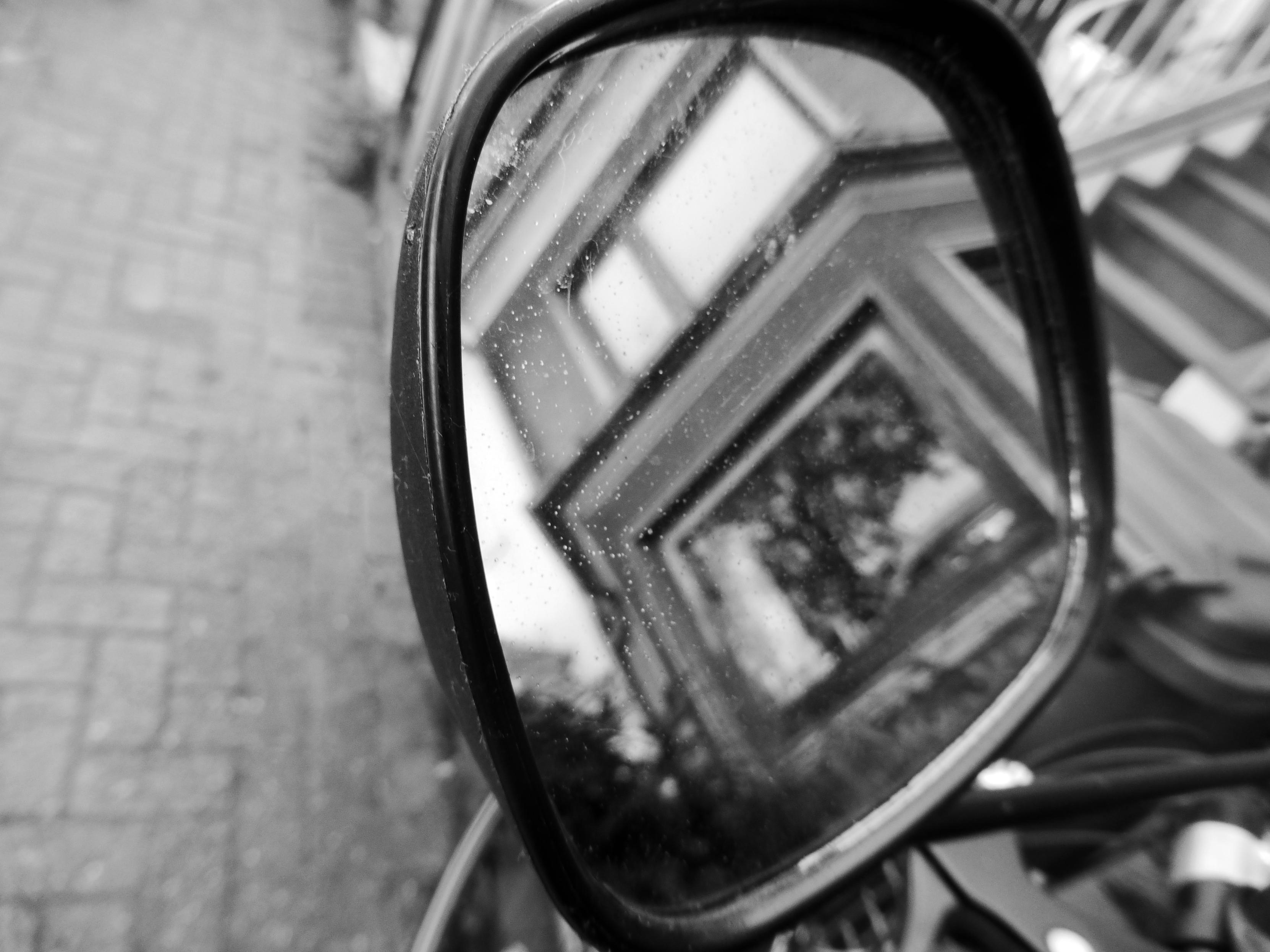 Black-and-white photograph of a motorcycle side mirror reflecting a building facade with several bright rectangular windows, geometric architectural lines, and blurred trees; a paved sidewalk appears beside the mirror.