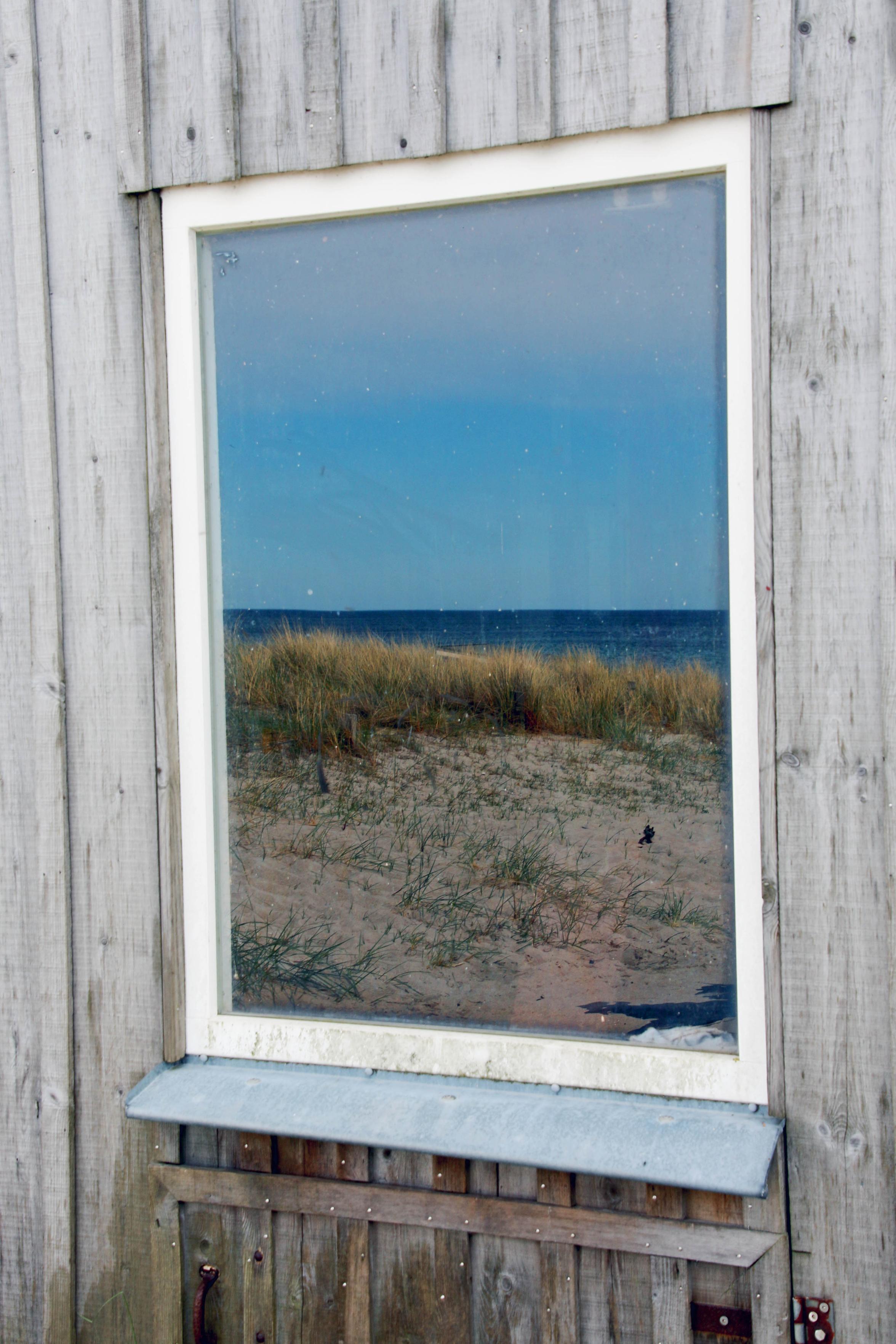 In dem Fenster einer grauen Strandhütte spiegelt sich ein blauer Himmel, darunter das Meer, Dünengras und Strand so perfekt, als schaue man direkt aus dem Fenster aufs Meer.

A blue sky is reflected in the window of a grey beach hut; beneath it, the sea, the dune grass and the beach are so perfectly captured that it is as if you were looking out of the window directly at the sea.