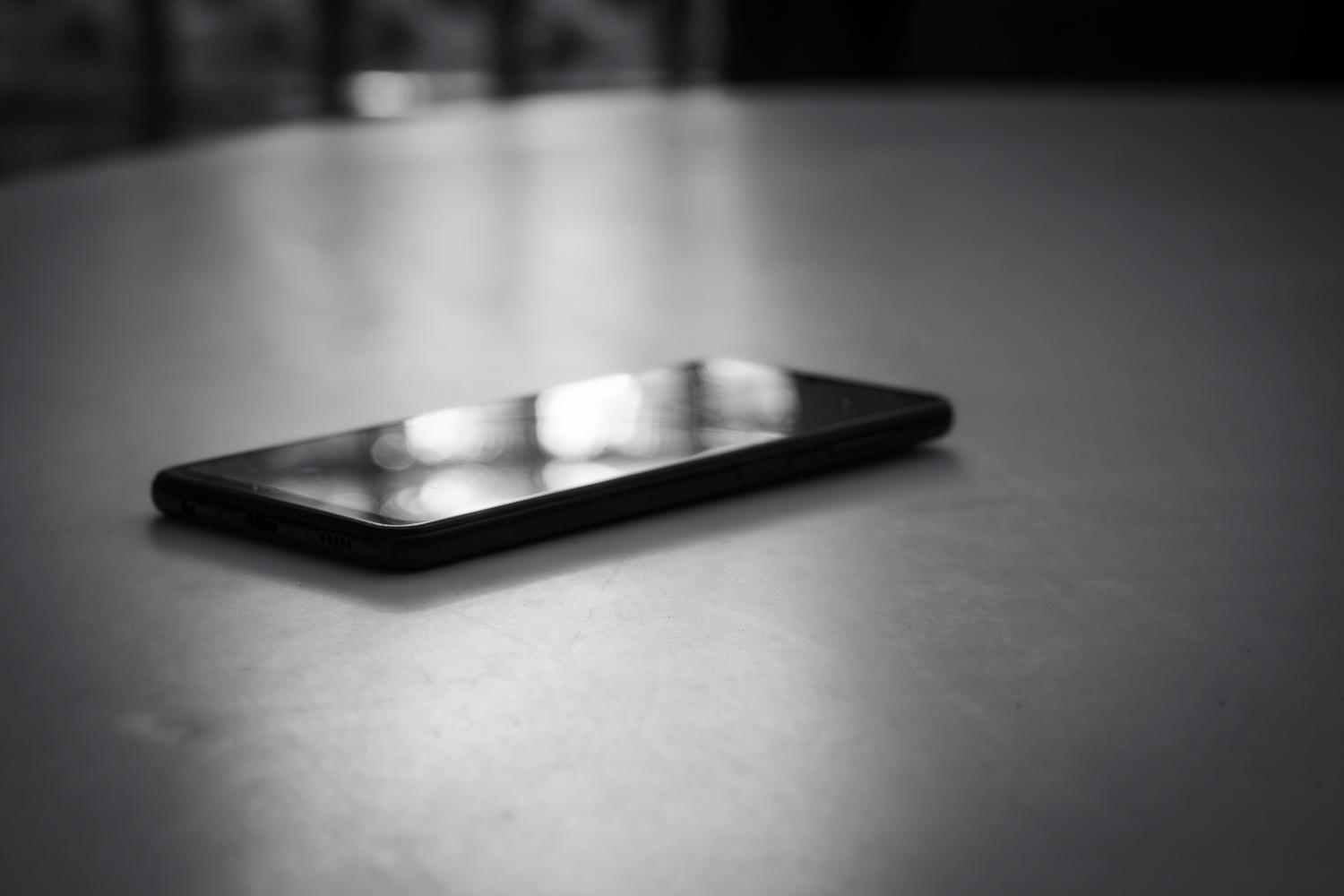 Schwarz-weisse Nahaufnahme eines Smartphones, das flach auf einer glatten Oberfläche liegt. Das Gerät ist leicht schräg von vorne zu sehen, während sich helle Lichtreflexe auf dem dunklen Bildschirm spiegeln. Der Hintergrund ist unscharf und dunkel, wodurch das Smartphone als zentrales Motiv hervorgehoben wird.