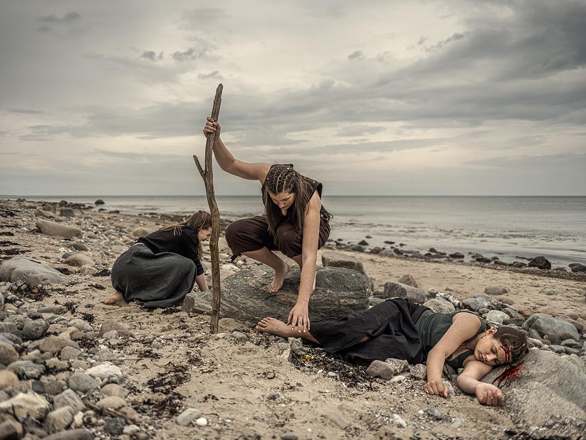 Szene aus einer Bildergeschichte. Drei Mädchen im Teenager-Alter am Strand. Sie sind barfuß, ihre Kleidung wirkt wie aus Piraten-Zeiten. Rechts im Bild liegt ein Mädchen mit einer blutenden Kopfwunde am Strand. Ihr Kopf liegt auf einem Stein, auf dem auch Blut zu sehen ist. In der Mitte hockt das älteste Mädchen auf einem Stein und beugt sich zu einem kleinen Säckchen mit Gold- und Silberschmuck. Etwas weiter hinten links im Bild erhebt sich ein anderes Mädchen gerade schreiend.