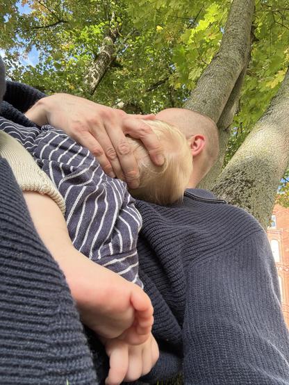 Ich lehne mit Baby an einem Baum. Das Baby schläft mit nackten Füßen. Man sieht seine Dichte Krone und im Hintergrund eine hohe Häuserwand aus Beckstein und blauer Himmel.