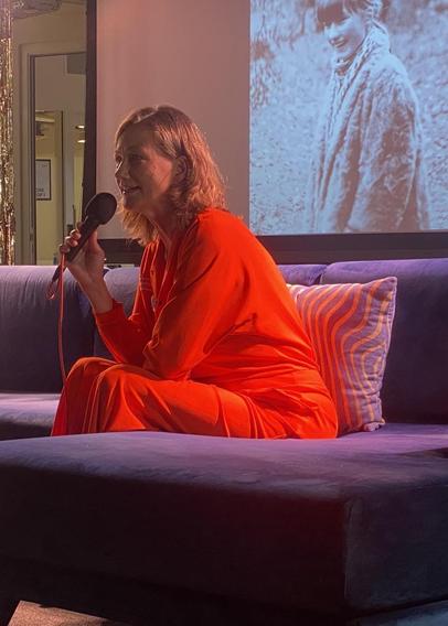 Silke in leuchtend hellrotem Overall auf einem dunklen Sofa. Dahinter eine Leinwand, auf der sie mit 13 zu sehen ist.