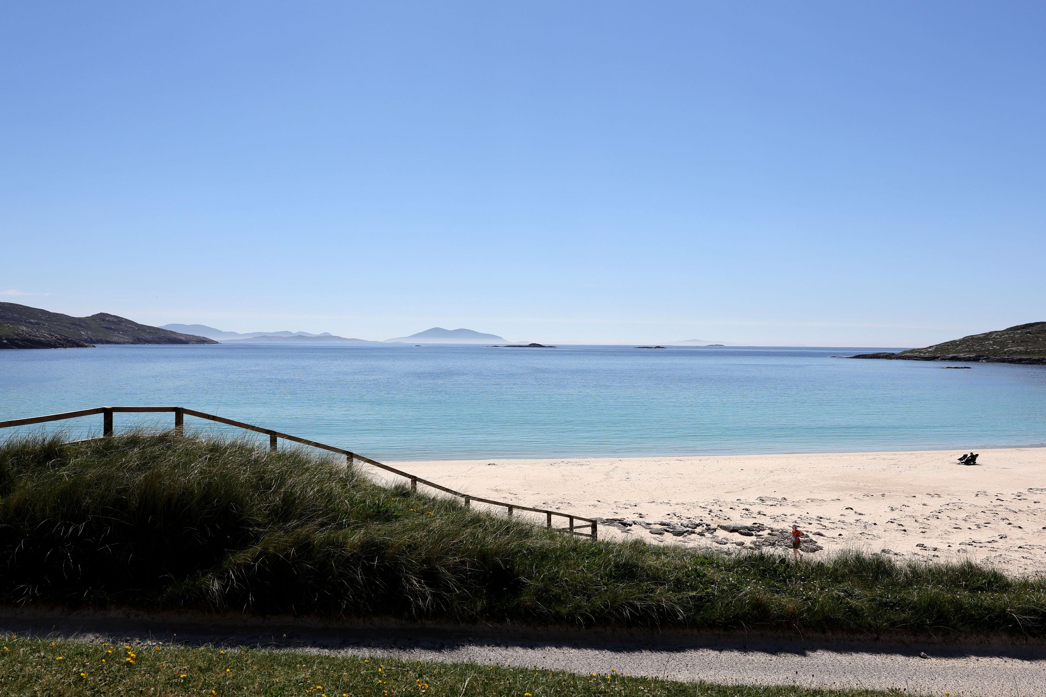Ein strahlend blauer Himmel, darunter türkisfarbenes Meer, weißer Sandstrand und eine hügelige Dünenlandschaft, eine Geländer aus Holz führt an den Strand an dem zwei Menschen sitzen. Im Hintergrund Berge und Hügel. (Mai 2025)

A bright blue sky, including turquoise sea, white sandy beach and a hilly dune landscape, a wooden railing leads to the beach where two people sit. In the background mountains and hills. (May 2025)