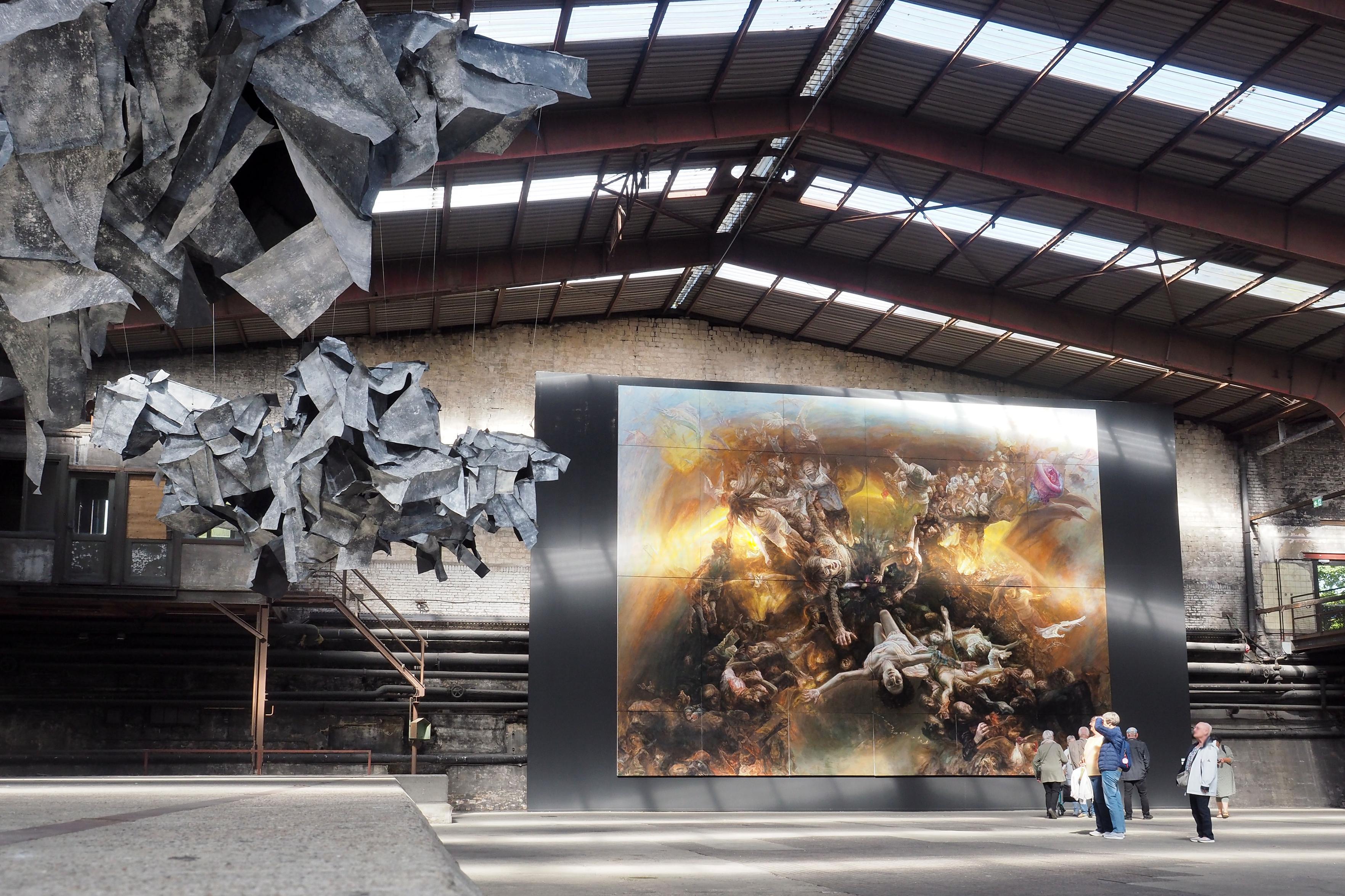 Zwei Kunstwerke in der Carlshütte in Büdelsdorf: Links hängen die Installlation "Exuvie" aus Papier der Künstlerin Anna Myga Kasten von der Decke, an der Rückwand der Halle ist das riesige Wandbild "Eye in the sky" des chinesischen Künstlers Liu Yaming zu sehen. Ein paar Menschen stehen vor den Werken und betrachten diese. 

Two works of art in the Carlshütte in Büdelsdorf: On the left hang the installation "Exuvie" made of paper by the artist Anna Myga Kasten from the ceiling, on the back wall of the hall is the huge mural "Eye in the sky" by the Chinese artist Liu Yaming. A few people stand before the works and look at them.