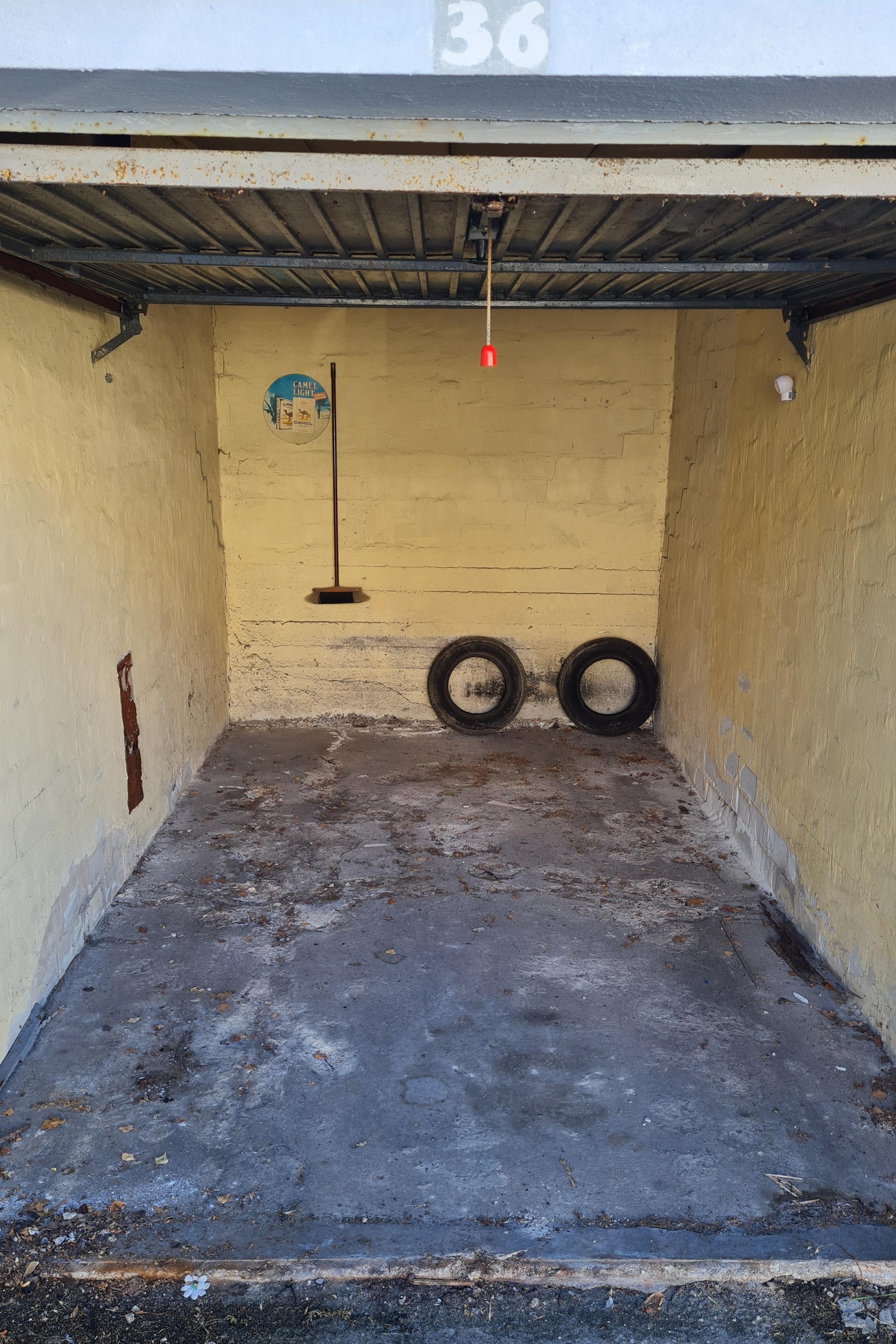 An abandoned yellow-painted garage with a corrugated metal roof. Two old tires rest against the back wall, while a broom hangs from the ceiling.