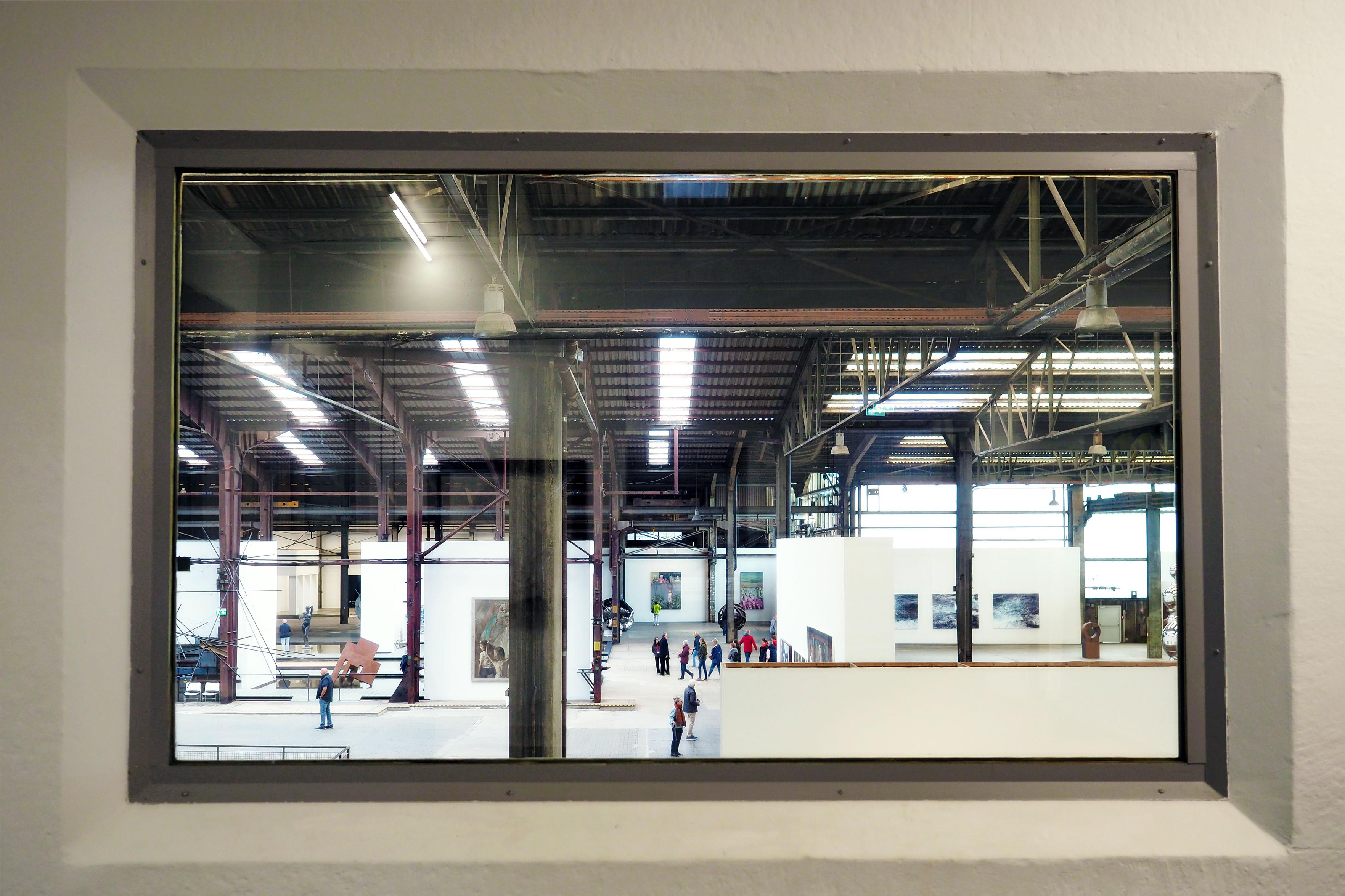 Blick aus einem Fenster von einem oberen Stockwerk auf eine Ausstellungshalle der Carlshütte in Büdelsdorf. Menschen gehen hin und her und betrachten Kunstwerke an weißen Wänden. Darüber Träger, Balken und Gestelle, die das Dach der Halle tragen sowie viele Rohre und Leuchtstoffröhren.  

View from a window from an upper floor to an exhibition hall of the Carlshütte in Büdelsdorf. People go back and forth and look at artworks on walls. Above it, beams and racks that carry the roof of the hall, as well as many pipes and fluorescent tubes.