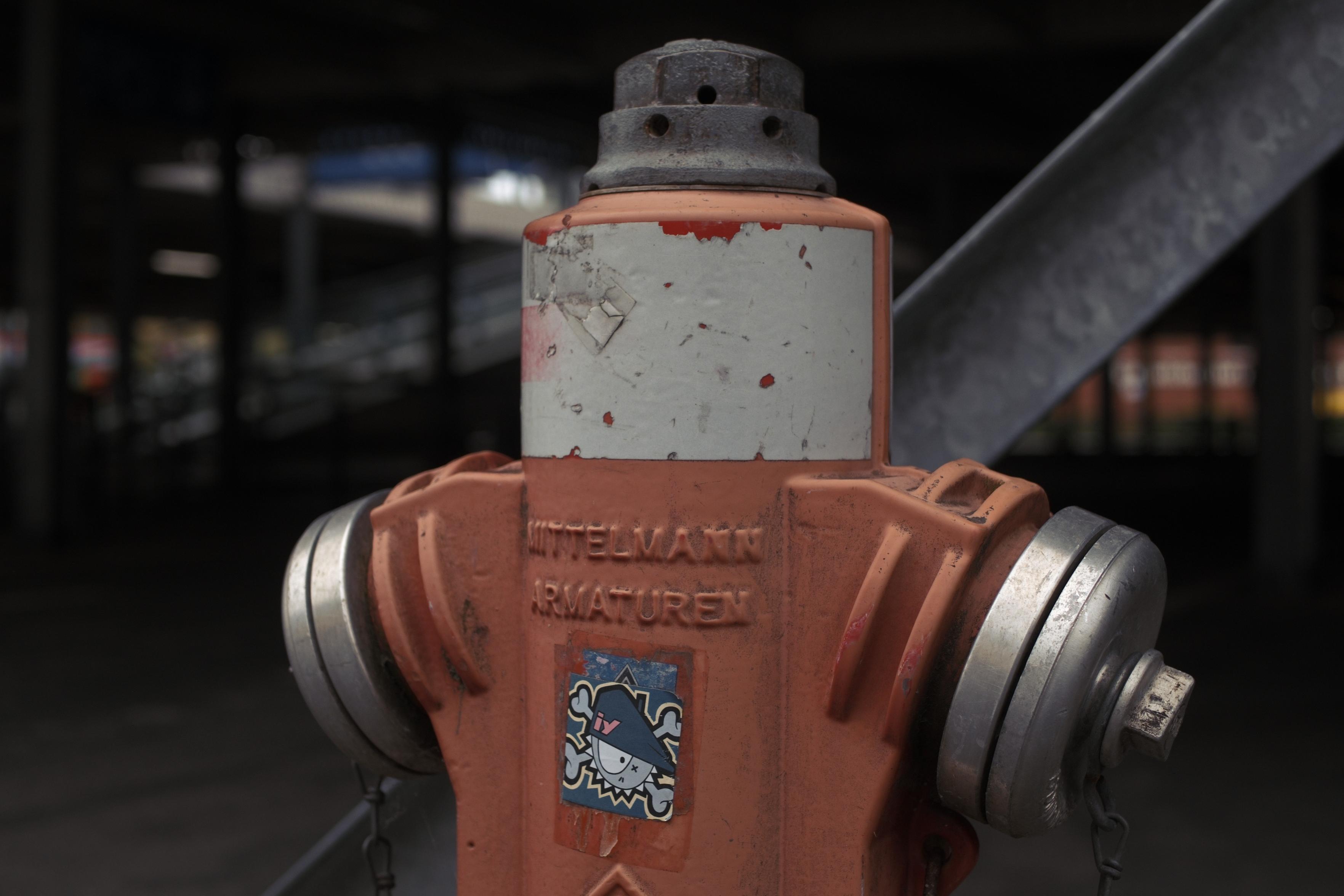 Deutsch:
Verwitterter roter Hydrant mit silbernen seitlichen Anschlüssen und einem schwarzen Rohr dahinter. On the front is a small sticker featuring a cartoon character.

English:
Weathered red hydrant with silver side connections and a black pipe behind it. On the front is a small sticker featuring a cartoon character.