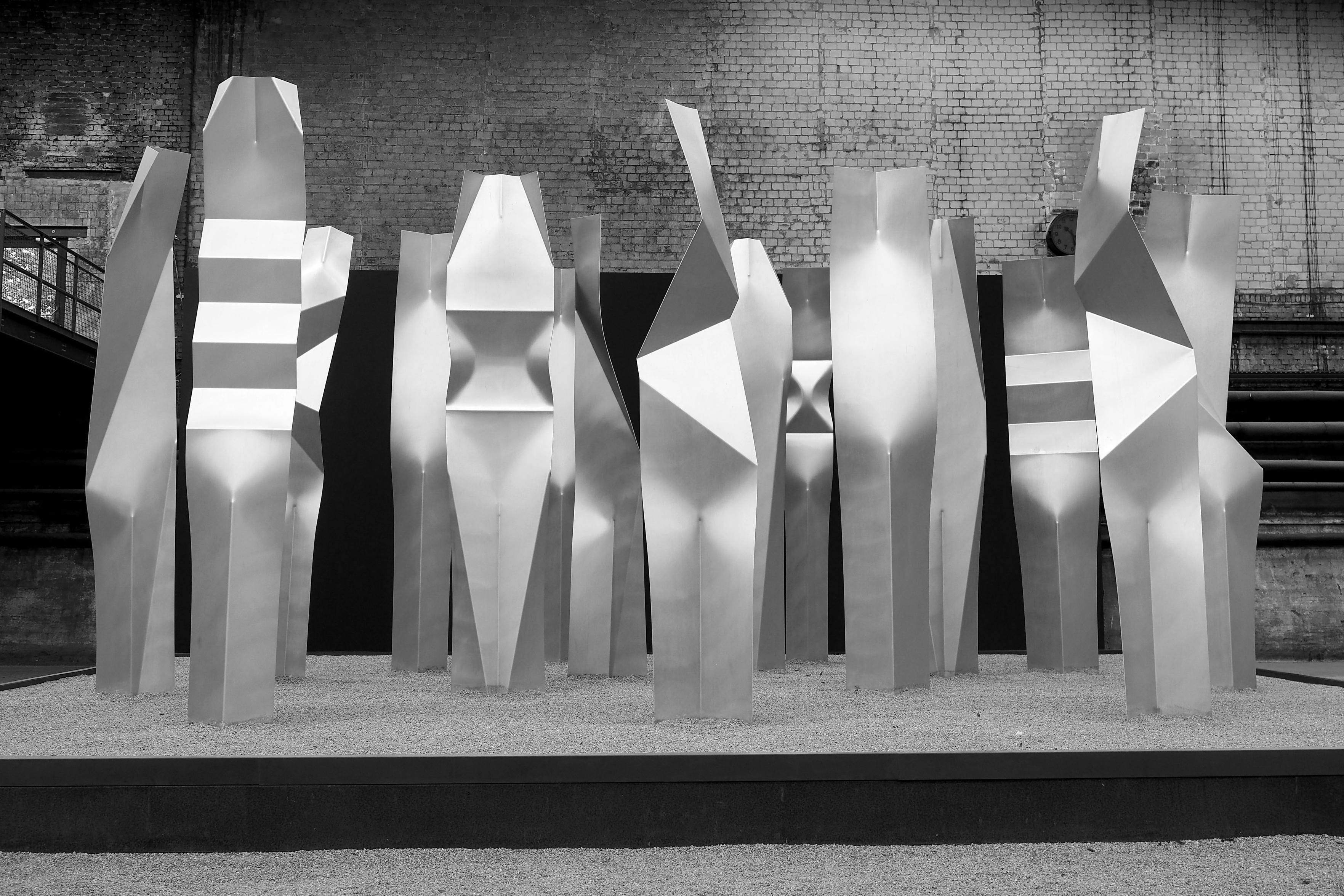 Schwarz-weiße Aufnahme von 15 großen Skulpturen aus Aluminium, die als Gruppe beieinander stehen. Die sehr abtrakten Formen wirken dennoch menschlich.

Black and white shot of 15 large sculptures made of aluminum, which stand together as a group. The very abstract forms nevertheless appear human.

NordArt 2026, Renes Krzystof - Universale 4000x5