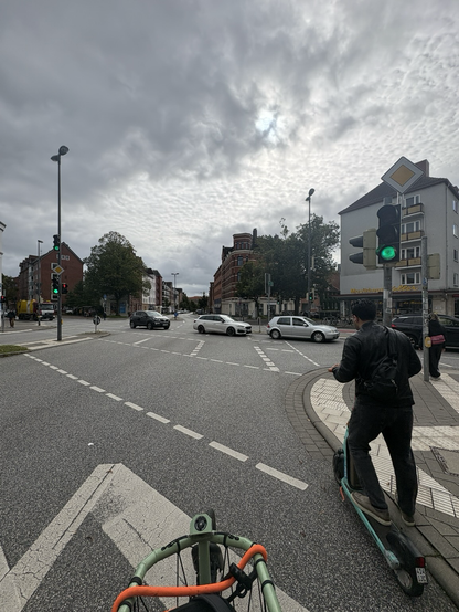 Foto einer Kreuzung vom Fahrrad in Fahrtrichtung geradeaus. Die Spiel zeigt grün. Drei Autofahrende von links kommend und nach rechts fahrend blockieren die Fahrbahn in Fahrtrichtung in kompletter Breite mit ihren Autos, weil sie in die Kreuzung einfuhren, obwohl die Ausfahrt durch Stau längst nicht mehr zu nutzen war.