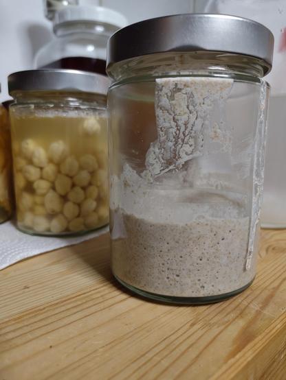 Im Vordergund ein Schraubglas mit aufgegangenem Roggensauerteig. Es steht auf einer Holzfläche. Auf einem Papier-Küchentuch dahinter sind weitere Fermente in Schraubläsern sichtbar, als nächstes rohe, fermentierte Kichererbsen und ein großes Gefäß mit einem dunkel-rotem Ferment 