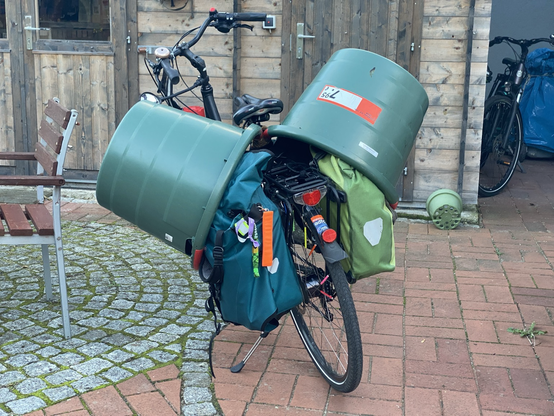 mein Rad von hinten. Es handelt sich um ein schwarzes TrekkingBike, hinter dem Sattel sind zwei riesige GartenTonnen links und rechts auf dem Fahrrad befestigt. darunter erkennt man links einen FahrradRucksack und rechts eine große OrtliebFahrradtasche