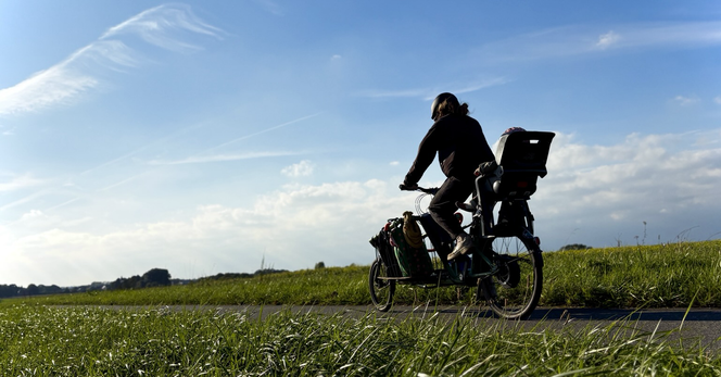 Radfahrerin mit Kind im Kindersitz über dem Hinterrad fährt entlang von Wiesen und Wäldern unter blauem Himmel